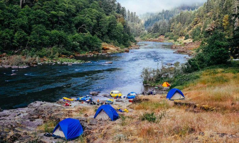 A scenic riverside campsite on the Rogue River featuring blue camping tents on a grassy bank and colorful rafts moored in the water.